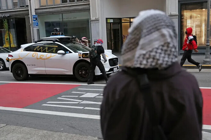 A protester graffitiing a Waymo car during the latest anti-ICE protest in downtown San Francisco. Mainstream media recorded Waymo vehicles spray-painted with slogans like “Fuck ICE” and “People Over Profit,” as well as red triangles marked in videos of Zionist soldiers in Palestinian resistance videos. Photo from Reddit